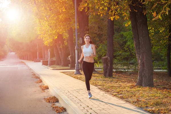 Jonge mooie vrolijke en sexy runner meisje met mooi lichaam in de herfst gekleurde park. met zon achtergrondverlichting is kijken camera met geluk en glimlach met witte sport pak. — Stockfoto