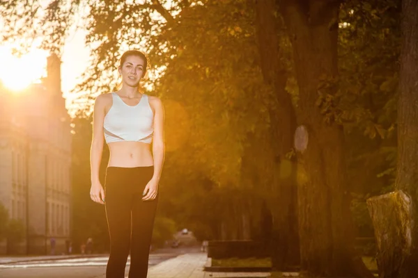 Jonge mooie vrolijke en sexy runner meisje met mooi lichaam in de herfst gekleurde park. met zon achtergrondverlichting is kijken camera met geluk en glimlach met witte sport pak. — Stockfoto