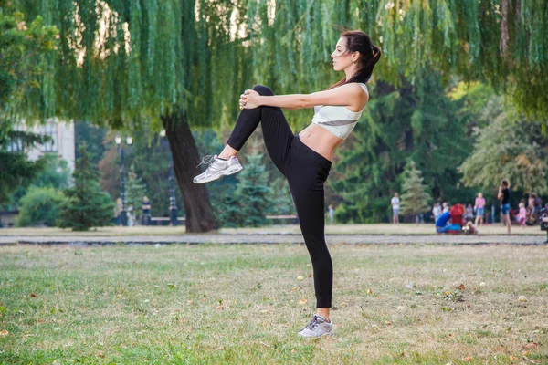 Young happy beautiful sexy girl is stretching in the park garden. people background. with dark hair and black and white sport suit and happiness and smile. on summer or autumn time season. — Stockfoto