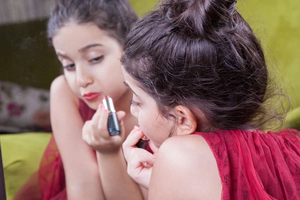 Pequeña hermosa chica árabe del medio oriente con bonito vestido rojo y labios haciendo maquillaje cuidadosamente en casa en el espejo. 8-10 años . — Foto de Stock