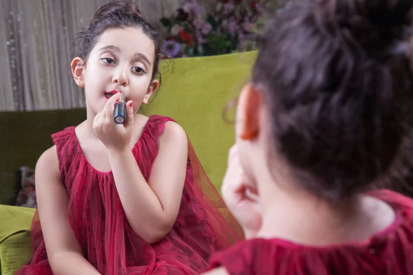 Pequena bela árabe menina do Oriente Médio com vestido vermelho bonito e lábios fazendo maquiagem cuidadosamente em casa no espelho. 8-10 anos . — Fotografia de Stock