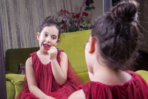 Small beautiful arab middle eastern girl with pretty red dress and lips doing makeup carefully at home in mirror. 8-10 years. — Stock Photo, Image