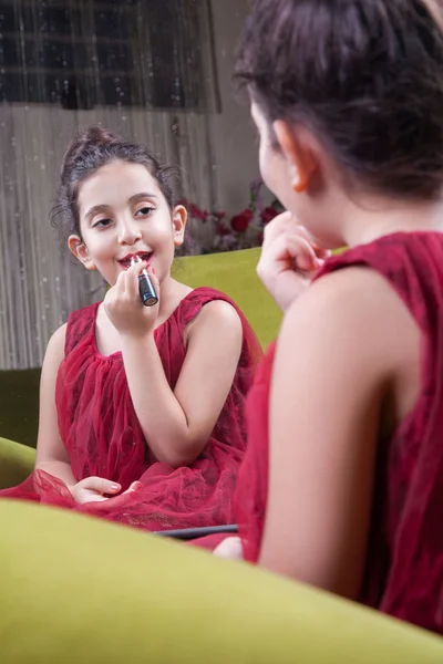 Pequeña hermosa chica árabe del medio oriente con bonito vestido rojo y labios haciendo maquillaje cuidadosamente en casa en el espejo. 8-10 años . —  Fotos de Stock