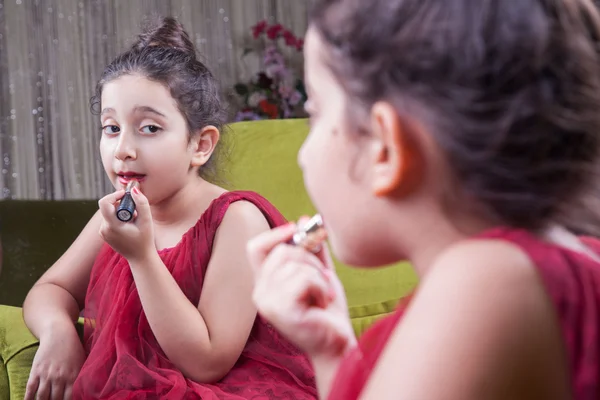 Pequeña hermosa chica árabe del medio oriente con bonito vestido rojo y labios haciendo maquillaje cuidadosamente en casa en el espejo. 8-10 años . —  Fotos de Stock