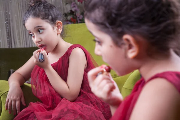 Pequeña hermosa chica árabe del medio oriente con bonito vestido rojo y labios haciendo maquillaje cuidadosamente en casa en el espejo. 8-10 años . —  Fotos de Stock
