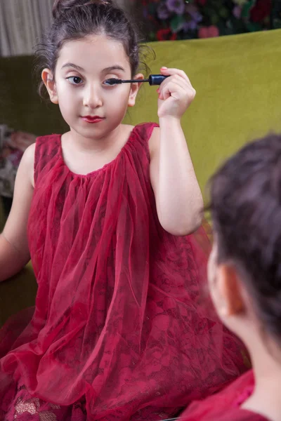 Μικρή όμορφη Άραβας μεσανατολική κορίτσι με όμορφο κόκκινο φόρεμα και χείλη κάνει μακιγιάζ, προσεκτικά στο σπίτι στον καθρέφτη. 8-10 χρόνια. — Φωτογραφία Αρχείου