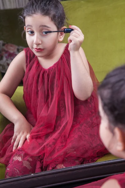 Pequeña hermosa chica árabe del medio oriente con bonito vestido rojo y labios haciendo maquillaje cuidadosamente en casa en el espejo. 8-10 años . — Foto de Stock