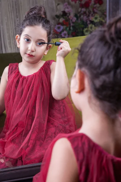 Pequeña hermosa chica árabe del medio oriente con bonito vestido rojo y labios haciendo maquillaje cuidadosamente en casa en el espejo. 8-10 años . — Foto de Stock