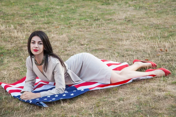 Mulher jovem sexy bonita com vestido clássico deitado na bandeira americana no parque. modelo de moda segurando-nos sorrindo e olhando para a câmera. Estilo de vida dos EUA com sorriso de dente . — Fotografia de Stock