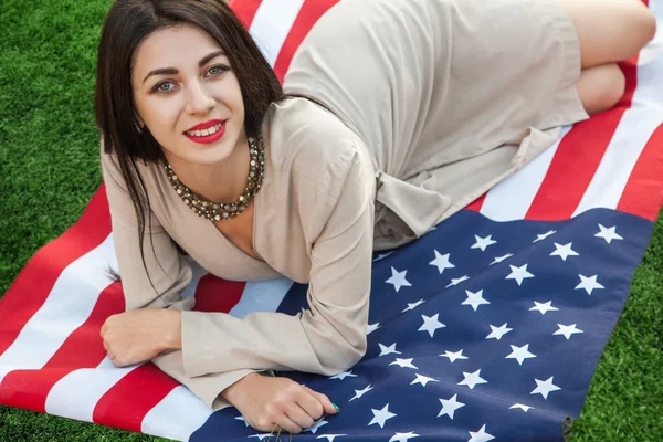 Mulher jovem sexy bonita com vestido clássico deitado na bandeira americana no parque. modelo de moda segurando-nos sorrindo e olhando para a câmera. Estilo de vida dos EUA com sorriso de dente . — Fotografia de Stock