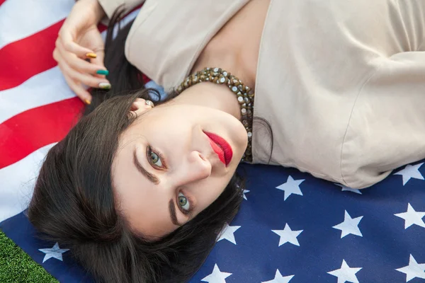 Mulher jovem sexy bonita com vestido clássico deitado na bandeira americana no parque. modelo de moda segurando-nos sorrindo e olhando para a câmera. Estilo de vida dos EUA com sorriso de dente . — Fotografia de Stock