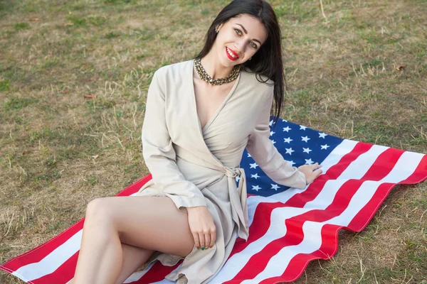 Piękna sexy młoda kobieta z klasyczna sukienka leżącej na amerykańską flagę w parku. modelka trzyma nas uśmiechając się i patrząc na kamery. USA styl życia uśmiech ząb. — Zdjęcie stockowe