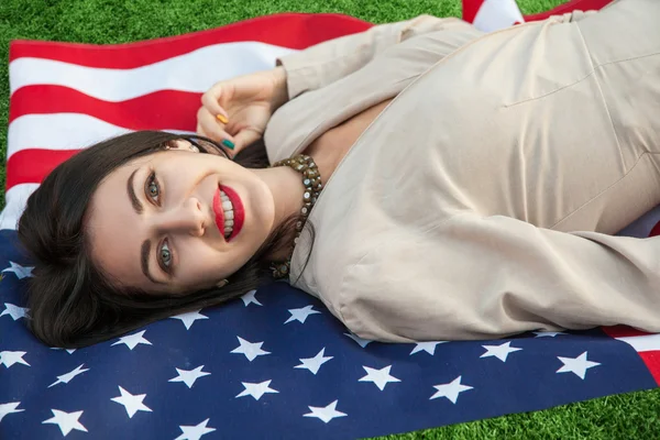 Красива сексуальна молода жінка з класичні сукні, лежачи на американський прапор в парку. фотомодель тримає нас, посміхаючись і, дивлячись на камеру. США стиль життя з зубастий посмішка. Стокове Зображення