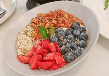 İçinde çilek, yaban mersini, muz, chia tohumu ve renkli bölümlerde sıralanmış çıtır yulaf bulunan taze meyve ve granola yoğurt kasesi. 