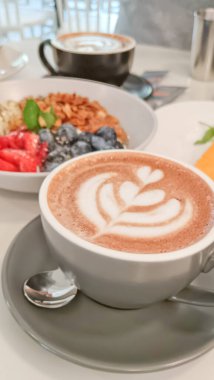 Sıcak bir fincan köpüklü latte renkli bir meyve ve granola kasesinin yanında, parlak bir kafe masasında.. 