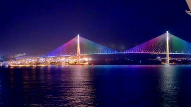 Busan, Güney Kore, 1 Ocak 2025, Busan, Güney Kore 'de bir asma kablo köprüsünün gece görüntüsü, gökkuşağı ışıklarıyla güzelce aydınlatılmış, suya ve deniz yolculuğundan gelen tekneye yansıyan.
