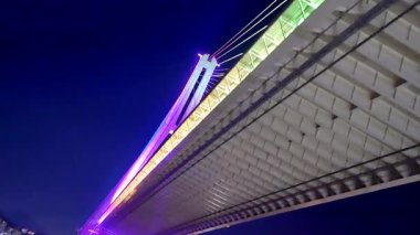 Busan, Güney Kore, 1 Ocak 2025, Busan, Güney Kore 'de bir asma kablo köprüsünün gece görüntüsü, gökkuşağı ışıklarıyla güzelce aydınlatılmış, suya ve deniz yolculuğundan gelen tekneye yansıyan.