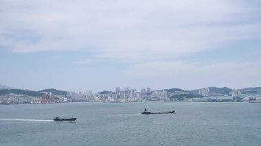 Kore, 1 Ocak 2025, geniş bir çekim Busan Limanı 'na yaklaşan büyük bir yolcu gemisi gösteriyor, sakin suları, liman tesisleri ve Busan, Güney ufuk çizgisi 