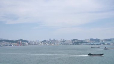 Kore, 1 Ocak 2025, geniş bir çekim Busan Limanı 'na yaklaşan büyük bir yolcu gemisi gösteriyor, sakin suları, liman tesisleri ve Busan, Güney ufuk çizgisi 