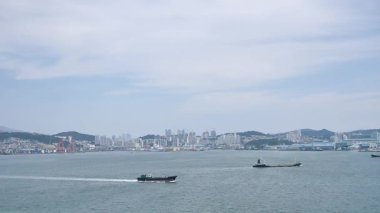 Kore, 1 Ocak 2025, geniş bir çekim Busan Limanı 'na yaklaşan büyük bir yolcu gemisi gösteriyor, sakin suları, liman tesisleri ve Busan, Güney ufuk çizgisi 