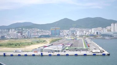Kore, 1 Ocak 2025, geniş bir çekim Busan Limanı 'na yaklaşan büyük bir yolcu gemisi gösteriyor, sakin suları, liman tesisleri ve Busan, Güney ufuk çizgisi 