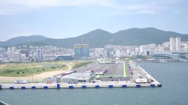 Kore, 1 Ocak 2025, geniş bir çekim Busan Limanı 'na yaklaşan büyük bir yolcu gemisi gösteriyor, sakin suları, liman tesisleri ve Busan, Güney ufuk çizgisi 