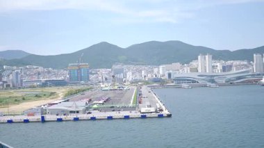 Kore, 1 Ocak 2025, geniş bir çekim Busan Limanı 'na yaklaşan büyük bir yolcu gemisi gösteriyor, sakin suları, liman tesisleri ve Busan, Güney ufuk çizgisi 