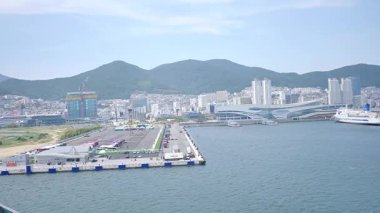 Kore, 1 Ocak 2025, geniş bir çekim Busan Limanı 'na yaklaşan büyük bir yolcu gemisi gösteriyor, sakin suları, liman tesisleri ve Busan, Güney ufuk çizgisi 