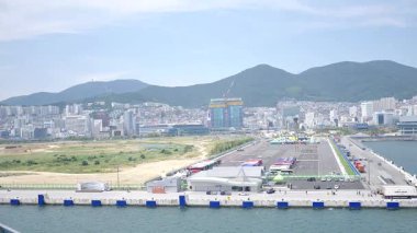 Kore, 1 Ocak 2025, geniş bir çekim Busan Limanı 'na yaklaşan büyük bir yolcu gemisi gösteriyor, sakin suları, liman tesisleri ve Busan, Güney ufuk çizgisi 