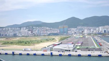 Kore, 1 Ocak 2025, geniş bir çekim Busan Limanı 'na yaklaşan büyük bir yolcu gemisi gösteriyor, sakin suları, liman tesisleri ve Busan, Güney ufuk çizgisi 