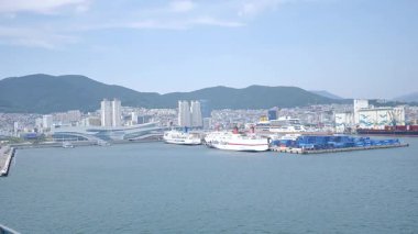 Busan, Güney Kore, 1 Ocak 2025 Geniş açılı çekimde Kore 'deki Busan limanına yaklaşan bir yolcu gemisi görülüyor. Kıyı kenti binaları, vinçler ve liman tesisleri görünüyor. 