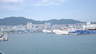 Busan, Güney Kore, 1 Ocak 2025 Geniş açılı çekimde Kore 'deki Busan limanına yaklaşan bir yolcu gemisi görülüyor. Kıyı kenti binaları, vinçler ve liman tesisleri görünüyor. 