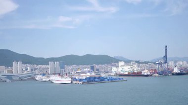 Busan, Güney Kore, 1 Ocak 2025 Geniş açılı çekimde Kore 'deki Busan limanına yaklaşan bir yolcu gemisi görülüyor. Kıyı kenti binaları, vinçler ve liman tesisleri görünüyor. 