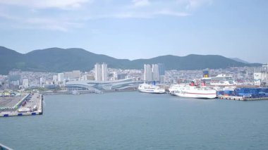 Busan, Güney Kore, 1 Ocak 2025 Geniş açılı çekimde Kore 'deki Busan limanına yaklaşan bir yolcu gemisi görülüyor. Kıyı kenti binaları, vinçler ve liman tesisleri görünüyor. 