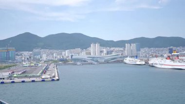 Busan, Güney Kore, 1 Ocak 2025 Geniş açılı çekimde Kore 'deki Busan limanına yaklaşan bir yolcu gemisi görülüyor. Kıyı kenti binaları, vinçler ve liman tesisleri görünüyor. 