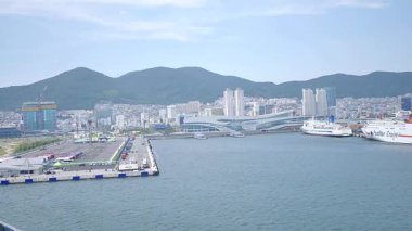 Busan, Güney Kore, 1 Ocak 2025 Geniş açılı çekimde Kore 'deki Busan limanına yaklaşan bir yolcu gemisi görülüyor. Kıyı kenti binaları, vinçler ve liman tesisleri görünüyor. 