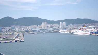 Busan, Güney Kore, 1 Ocak 2025 Geniş açılı çekimde Kore 'deki Busan limanına yaklaşan bir yolcu gemisi görülüyor. Kıyı kenti binaları, vinçler ve liman tesisleri görünüyor. 