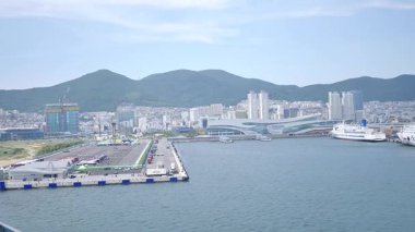 Busan, Güney Kore, 1 Ocak 2025 Geniş açılı çekimde Kore 'deki Busan limanına yaklaşan bir yolcu gemisi görülüyor. Kıyı kenti binaları, vinçler ve liman tesisleri görünüyor. 