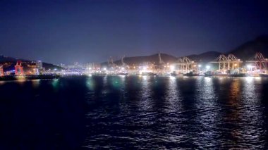 Geniş açı, Kore 'deki Busan Limanı' na gece yaklaşan bir yolcu gemisinin sahil kenti binaları, vinçler ve liman tesisleriyle birlikte görüldüğünü gösteriyor. 