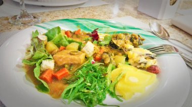 Beyaz tabakta canlı bir yahni, taze salata, kızarmış ekmek, yosun ve turşulu sebze karışımı bulunur.