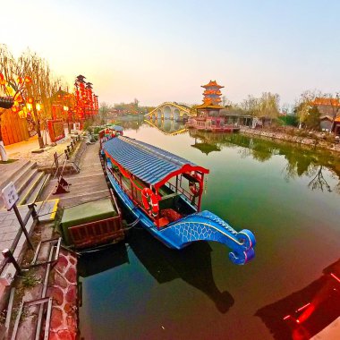 KaiFeng, Çin, 1 Ocak 2026, geleneksel Çin şehrinde panoramik nehir sahnesi, kırmızı botlar, ahşap pavyonlar, durgun su ve temiz gökyüzü altında tarihi binalar.