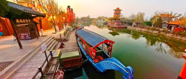 KaiFeng, Çin, 1 Ocak 2026, geleneksel Çin şehrinde panoramik nehir sahnesi, kırmızı botlar, ahşap pavyonlar, durgun su ve temiz gökyüzü altında tarihi binalar.