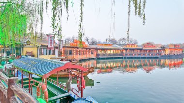 KaiFeng, Çin, 1 Ocak 2026, geleneksel Çin şehrinde panoramik nehir sahnesi, kırmızı botlar, ahşap pavyonlar, durgun su ve temiz gökyüzü altında tarihi binalar.