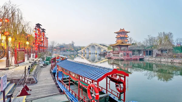 KaiFeng, Çin, 1 Ocak 2026, geleneksel Çin şehrinde panoramik nehir sahnesi, kırmızı botlar, ahşap pavyonlar, durgun su ve temiz gökyüzü altında tarihi binalar.