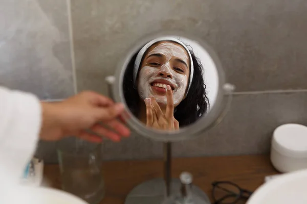 Gemischte Rassenfrau spiegelt sich im Spiegel, der im Badezimmer Gesichtscreme aufträgt. Selbstisolation zu Hause während der Covid 19 Coronavirus-Pandemie. — Stockfoto