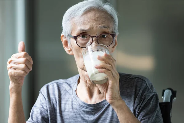 Asian senior woman drinking fresh milk from the glass,old elderly holding glass of milk,showing thumb up while drinking and looking at camera,health care,healthy nutrition,food and drinks concept