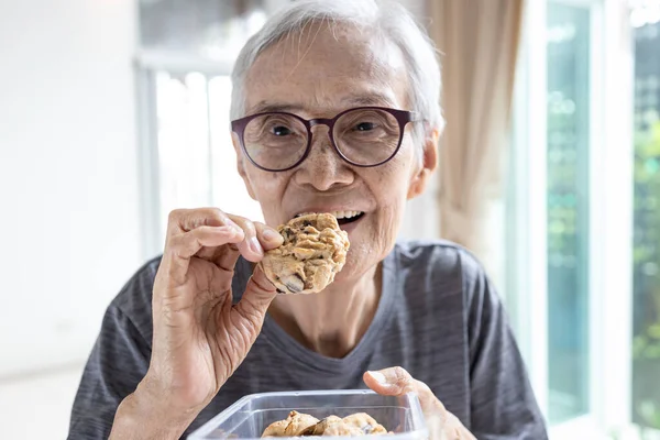 Happy asian senior woman eating chocolate chip cookie or tasting sweet cookie,natural tasty food,delicious snack,old elderly holding a box of cashew nut biscuits in her hand,breakfast in the morning