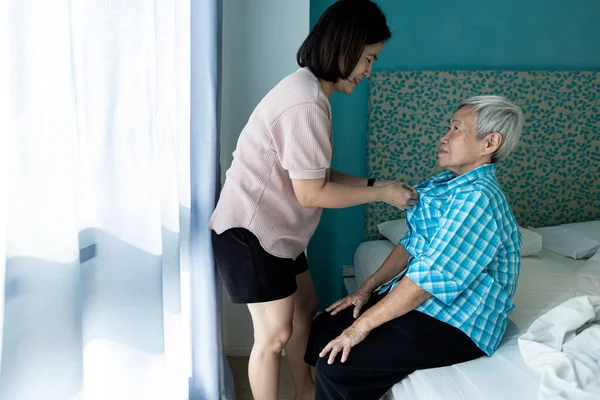 Asian female caregiver taking care of helping elderly patient get dressed,wearing clothes,button up on the shirt for happy senior grandmother,look after old people,support,daily life in a nursing home