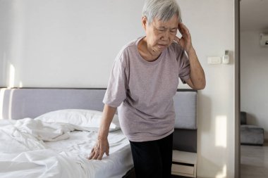 Baş dönmesinden muzdarip hasta Asyalı son sınıf öğrencisi, etrafında dönme hissi, dengesini kaybetme, yaşlı bir kadının yataktan kalktıktan sonra gözleri kapalı şakağına dokunması, baş dönmesi.