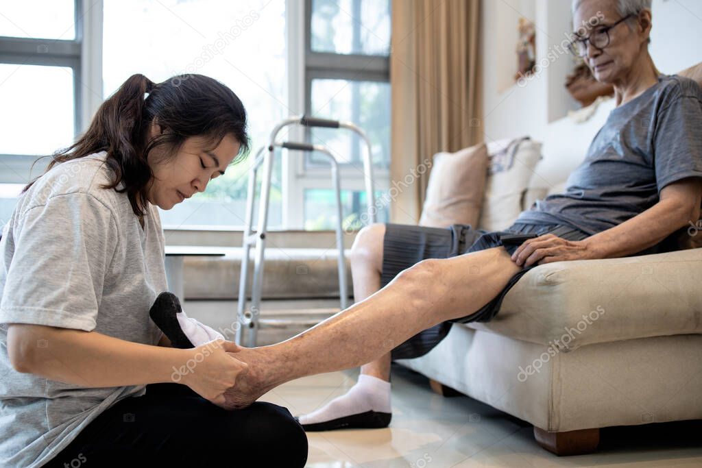 Asian daughter taking care of helping elderly,woman wear socks on feet of her senior mother,old people with cold toes,numb in foot,sensitivity to cold,putting on socks to warm up,health care at home
